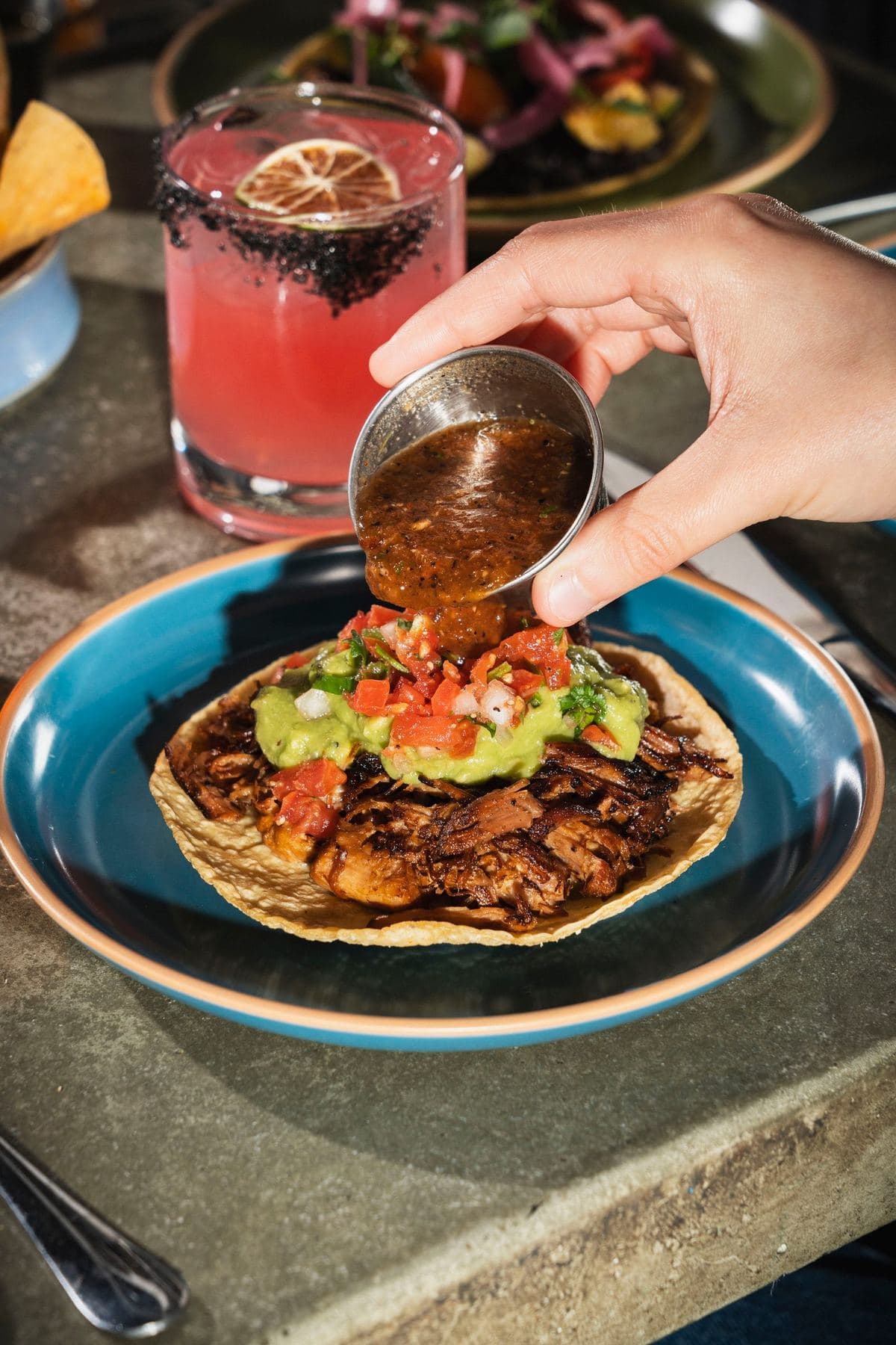 Pouring salsa over a taco with a pink cocktail in the background